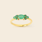 A gold ring featuring a central oval green emerald-cut stone surrounded by four smaller round green gemstones, set on a smooth gold band against a light beige background.