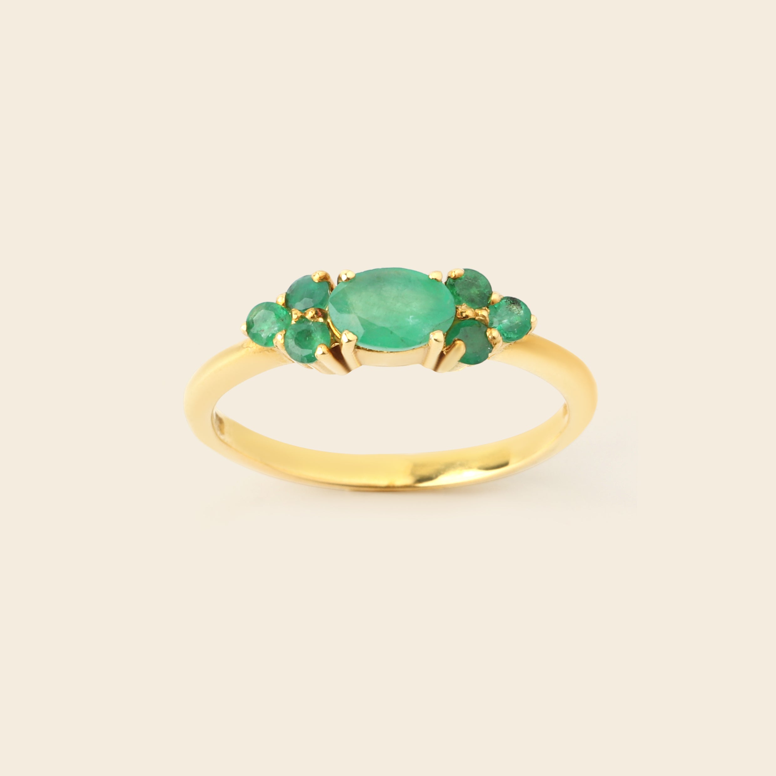 A gold ring featuring a central oval green emerald-cut stone surrounded by four smaller round green gemstones, set on a smooth gold band against a light beige background.