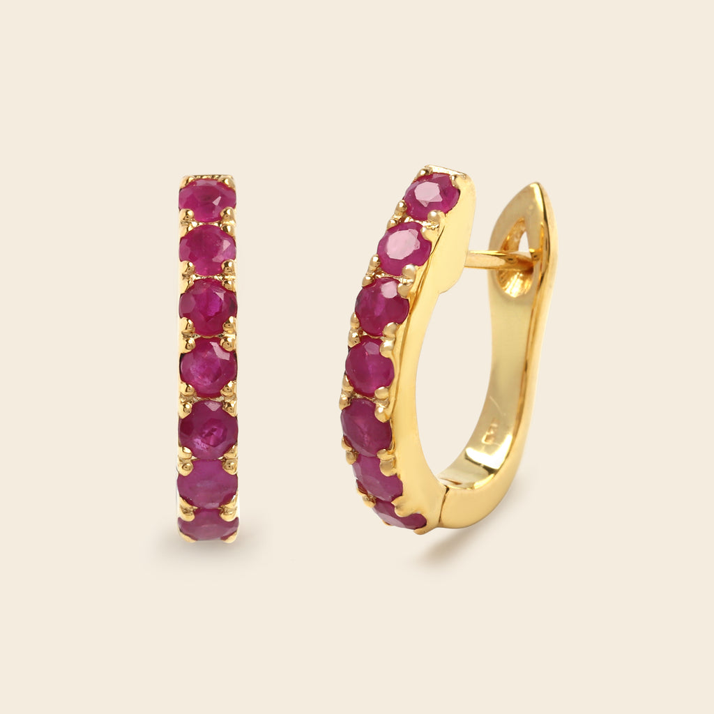 A pair of gold huggie hoop earrings with a row of round-cut pinkish-red gemstones, shown on a light beige background.
