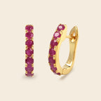 A pair of gold huggie hoop earrings with a row of round-cut pinkish-red gemstones, shown on a light beige background.