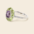 A silver ring featuring a floral cluster setting with a central oval purple amethyst gemstone surrounded by marquise-cut light green peridot gemstones and small clear accent stones, shown on a solid light beige background.