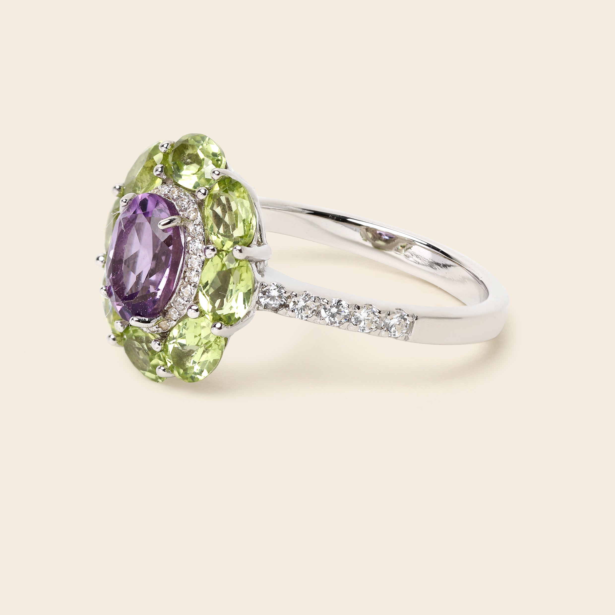 A silver ring featuring a floral cluster setting with a central oval purple amethyst gemstone surrounded by marquise-cut light green peridot gemstones and small clear accent stones, shown on a solid light beige background.