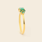 A yellow gold ring featuring a cluster of three oval-cut green gemstones, set against a solid, warm off-white background.