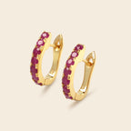 A pair of gold huggie hoop earrings embellished with a row of round-cut deep pinkish-purple gemstones, set against a solid light beige background.