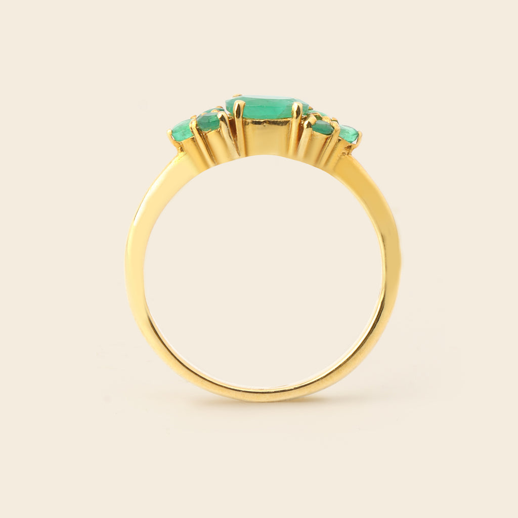 A gold ring featuring a central rectangular emerald-cut green gemstone flanked by three smaller round green gemstones, set against a plain light beige background.