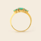 A gold ring featuring a central rectangular emerald-cut green gemstone flanked by three smaller round green gemstones, set against a plain light beige background.