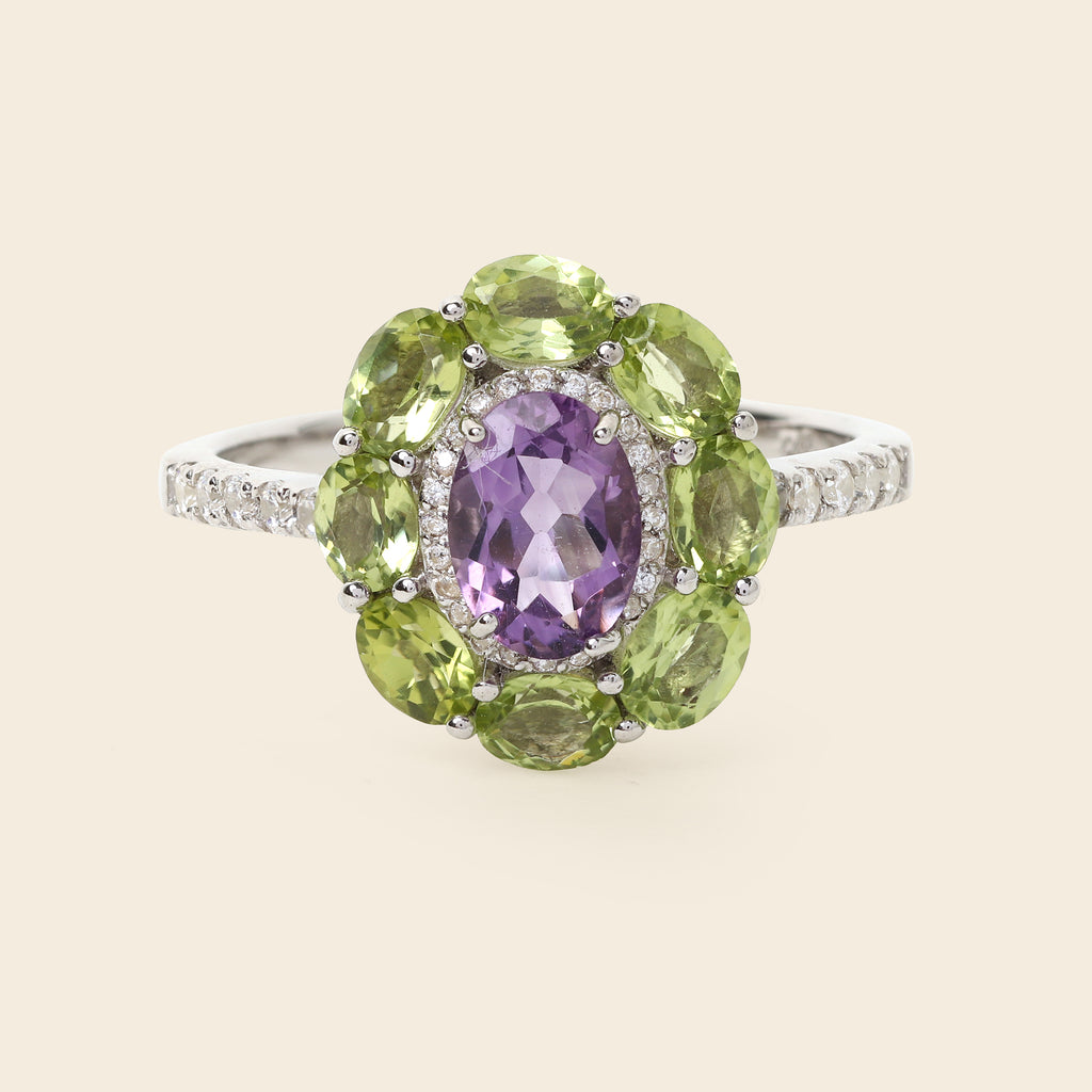A silver ring featuring a central oval purple amethyst gemstone surrounded by a halo of round green peridot gemstones, with small pavé-set clear stones along the band, shown on a light beige background.