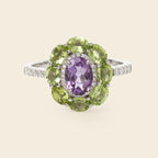 A silver ring featuring a central oval purple amethyst gemstone surrounded by a halo of round green peridot gemstones, with small pavé-set clear stones along the band, shown on a light beige background.