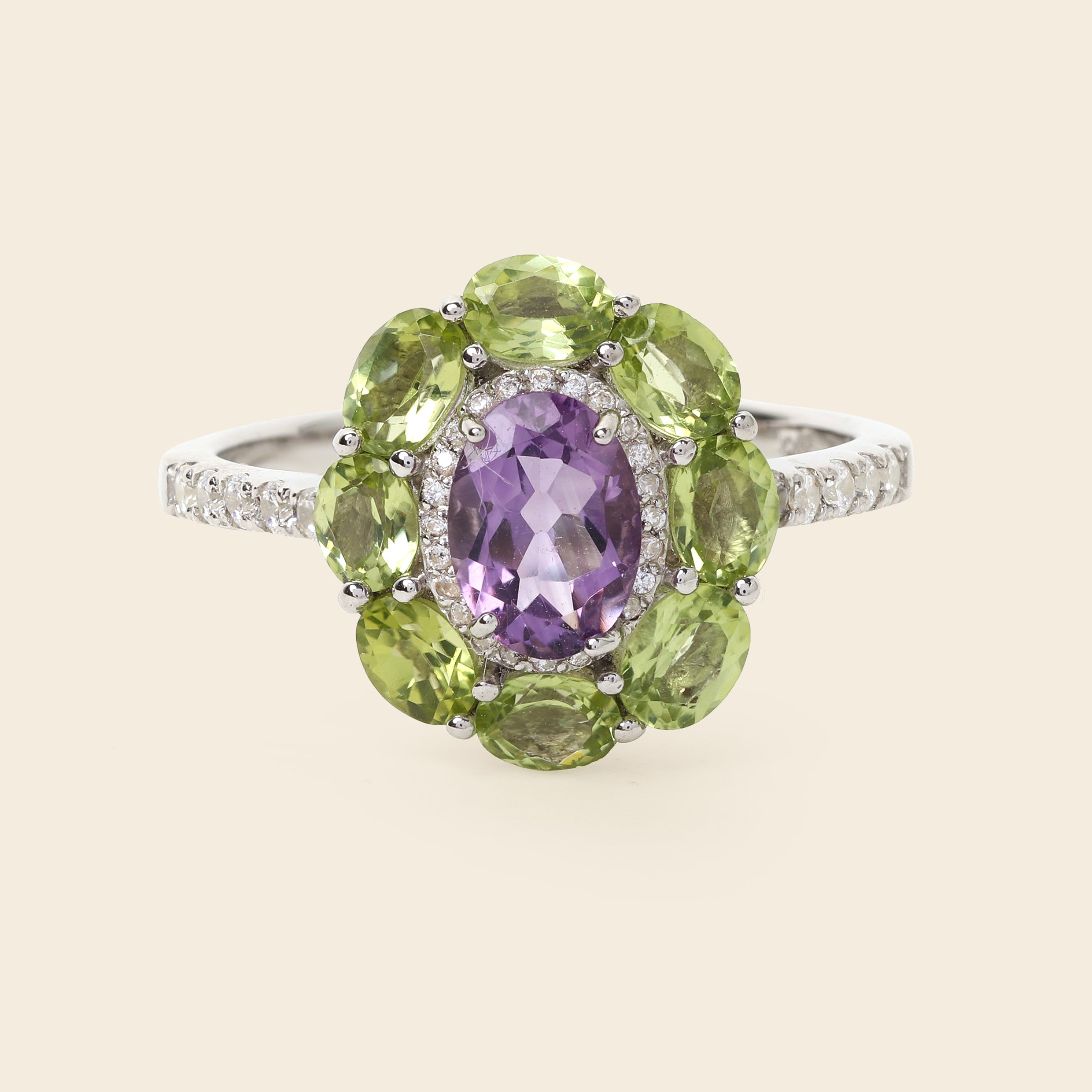 A silver ring featuring a central oval purple amethyst gemstone surrounded by a halo of round green peridot gemstones, with small pavé-set clear stones along the band, shown on a light beige background.
