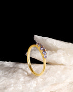 A gold ring with a curved row of small purple and blue gemstones, displayed on a textured white stone against a solid black background.