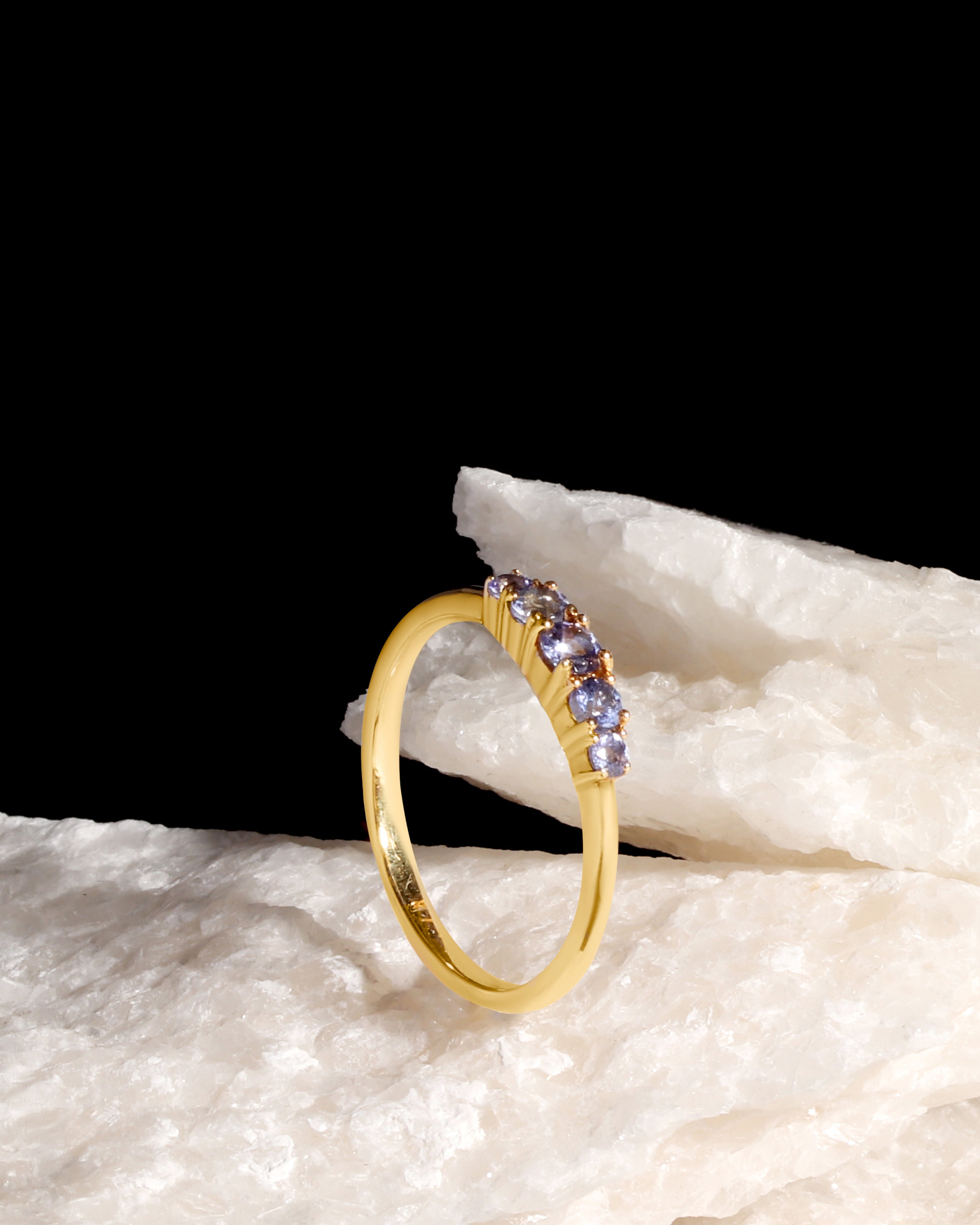 A gold ring with a curved row of small purple and blue gemstones, displayed on a textured white stone against a solid black background.