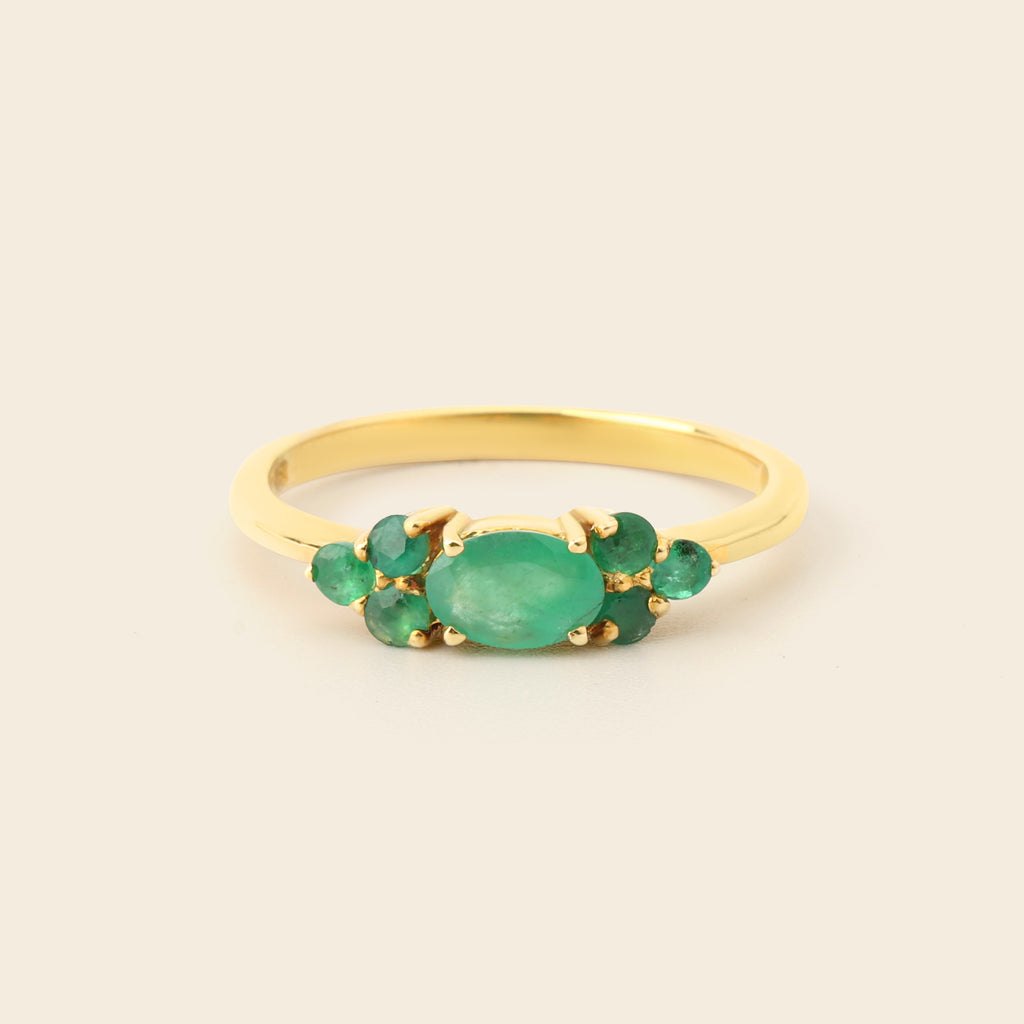 A gold ring featuring a central oval green emerald stone flanked by smaller round green emerald stones, set against a plain light beige background.
