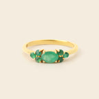 A gold ring featuring a central oval green emerald stone flanked by smaller round green emerald stones, set against a plain light beige background.