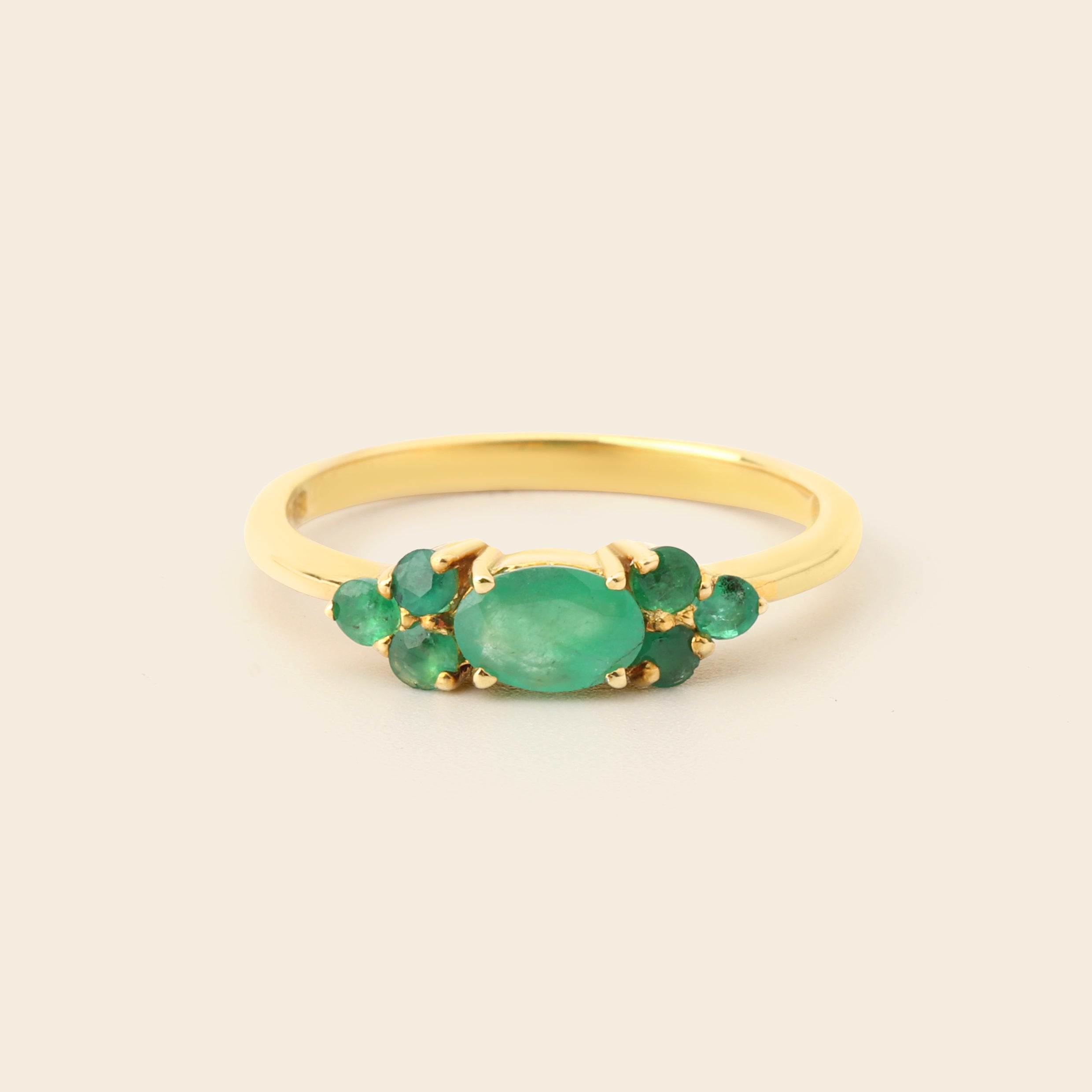 A gold ring featuring a central oval green emerald stone flanked by smaller round green emerald stones, set against a plain light beige background.