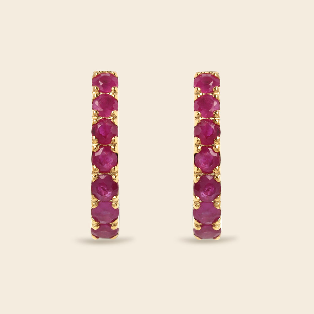 A pair of gold hoop earrings with a vertical channel setting of round-cut deep pinkish-red gemstones, set against a solid light beige background.