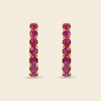 A pair of gold hoop earrings with a vertical channel setting of round-cut deep pinkish-red gemstones, set against a solid light beige background.