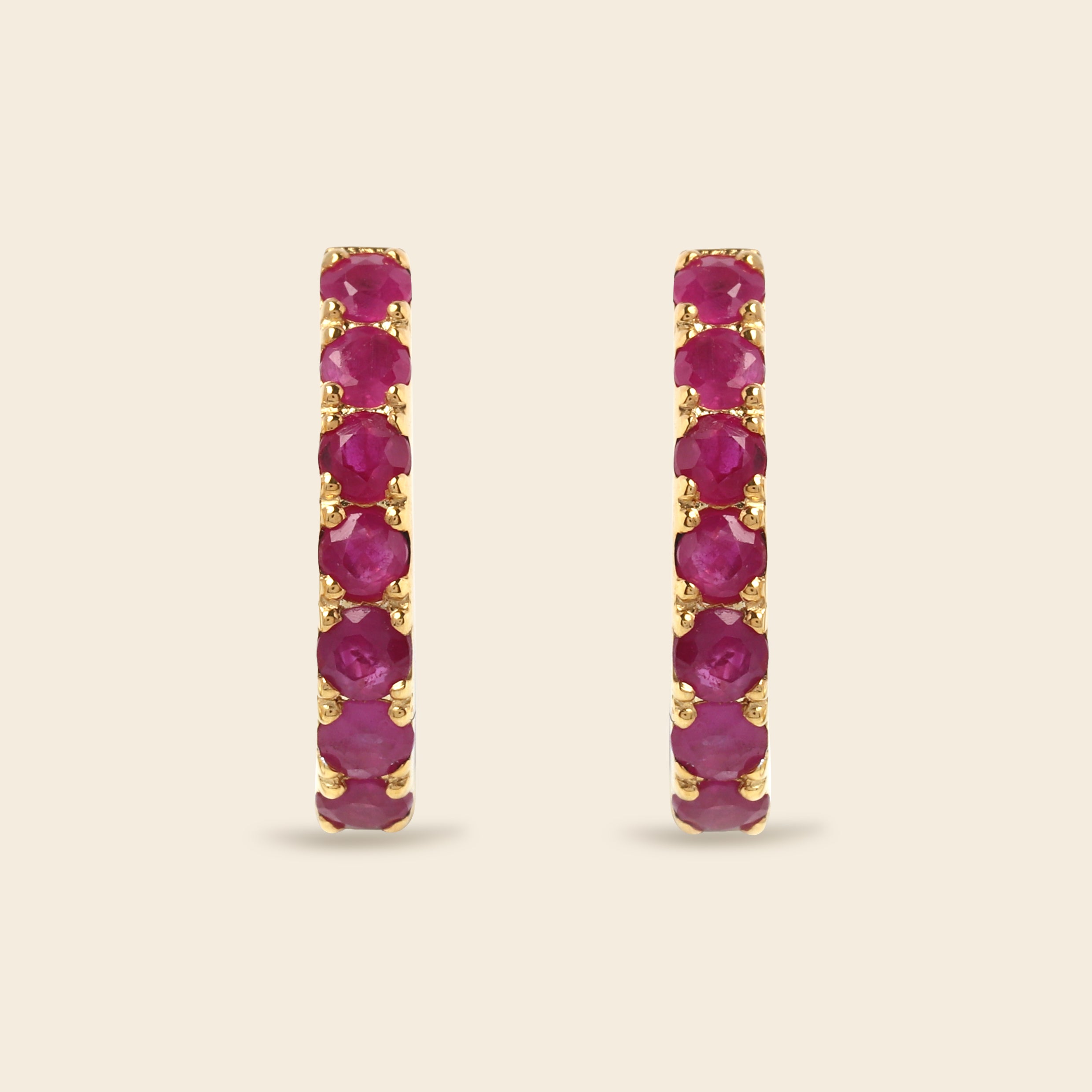 A pair of gold hoop earrings with a vertical channel setting of round-cut deep pinkish-red gemstones, set against a solid light beige background.