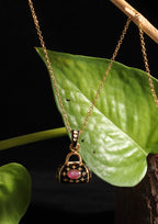 A gold pendant necklace featuring a miniature handbag charm with a pink gemstone and gold star details, hanging against a black background with a green leaf prop.