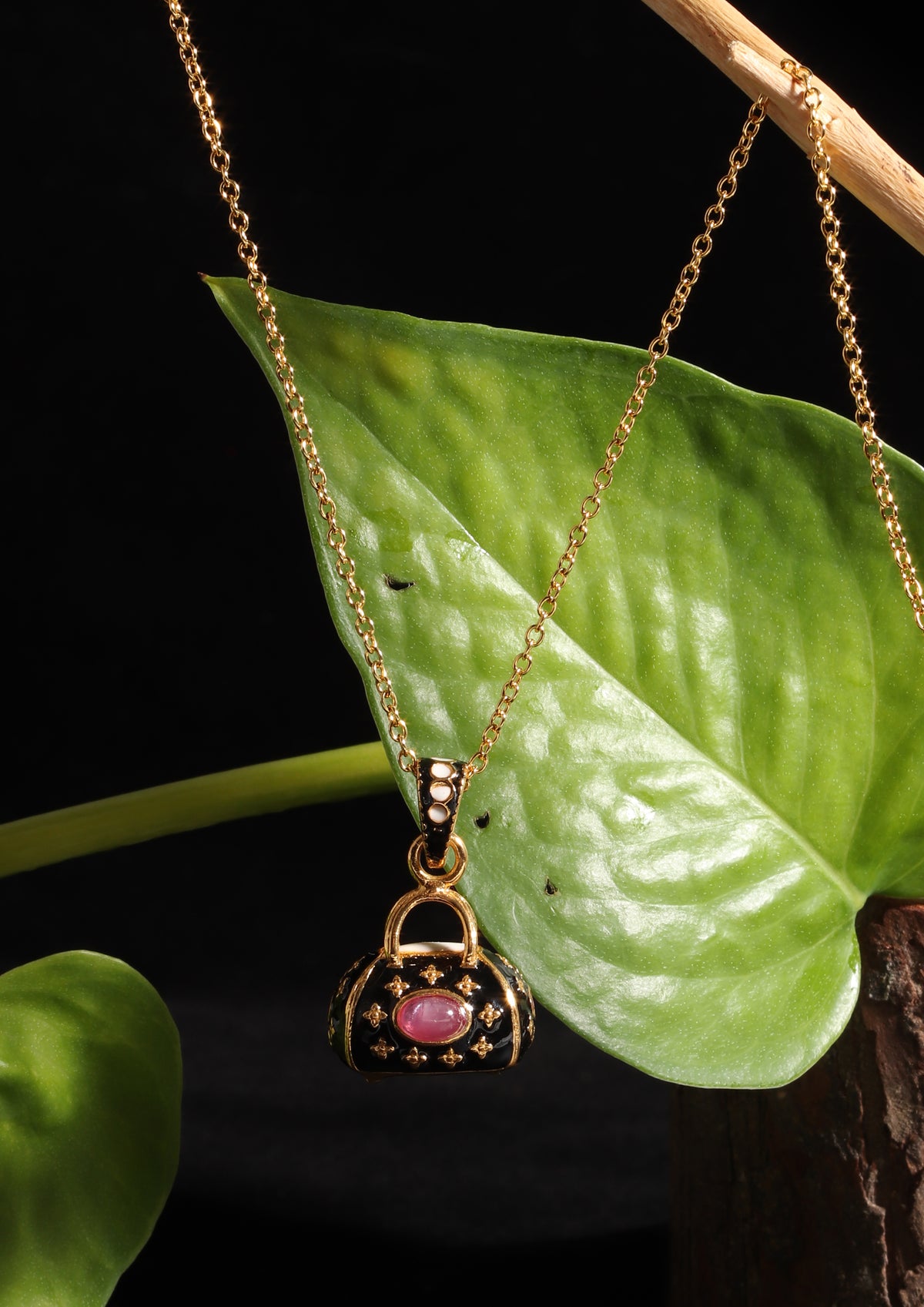 A gold pendant necklace featuring a miniature handbag charm with a pink gemstone and gold star details, hanging against a black background with a green leaf prop.