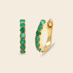 A pair of gold huggie hoop earrings with a row of round green emerald-cut gemstones, shown on a light beige background.