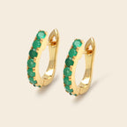 A pair of gold huggie hoop earrings embellished with a row of round green gemstones, shown on a light beige background.