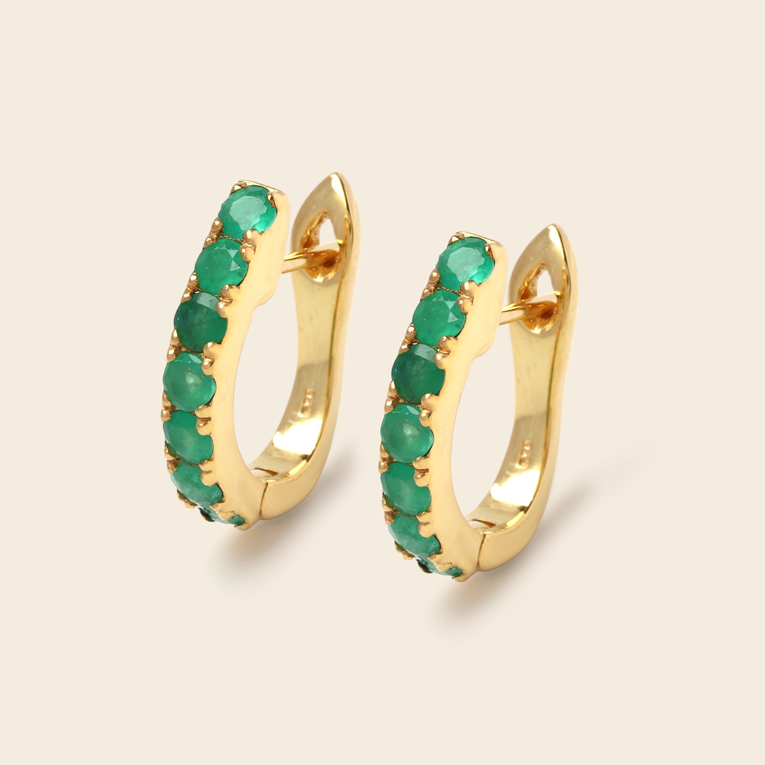 A pair of gold huggie hoop earrings embellished with a row of round green gemstones, shown on a light beige background.