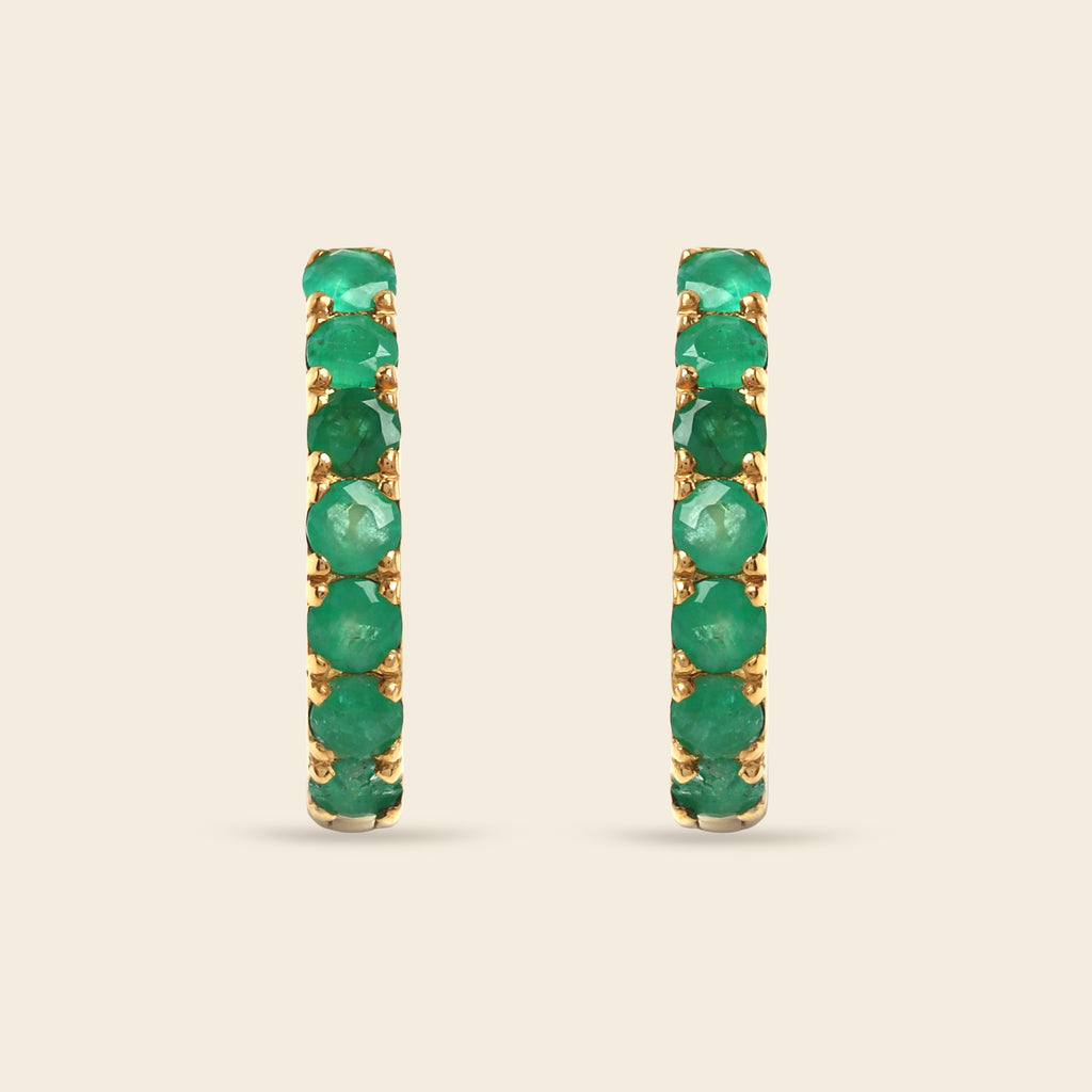 A pair of gold hoop earrings set with a full row of round green emeralds, shown against a solid light beige background.