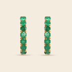 A pair of gold hoop earrings set with a full row of round green emeralds, shown against a solid light beige background.