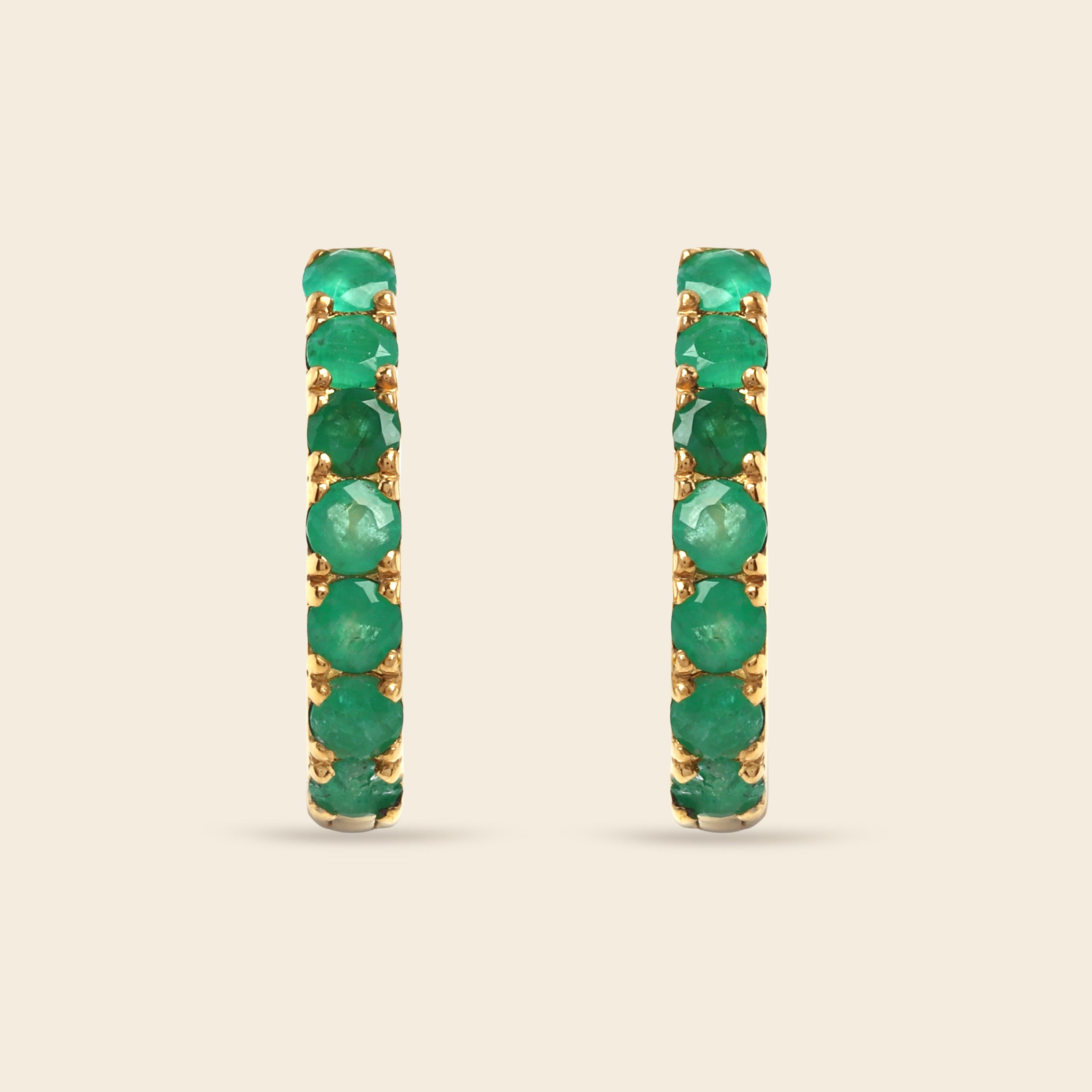 A pair of gold hoop earrings set with a full row of round green emeralds, shown against a solid light beige background.