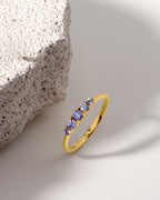 A gold ring with three round-cut blue gemstones, styled on a clean white surface next to a textured stone prop.
