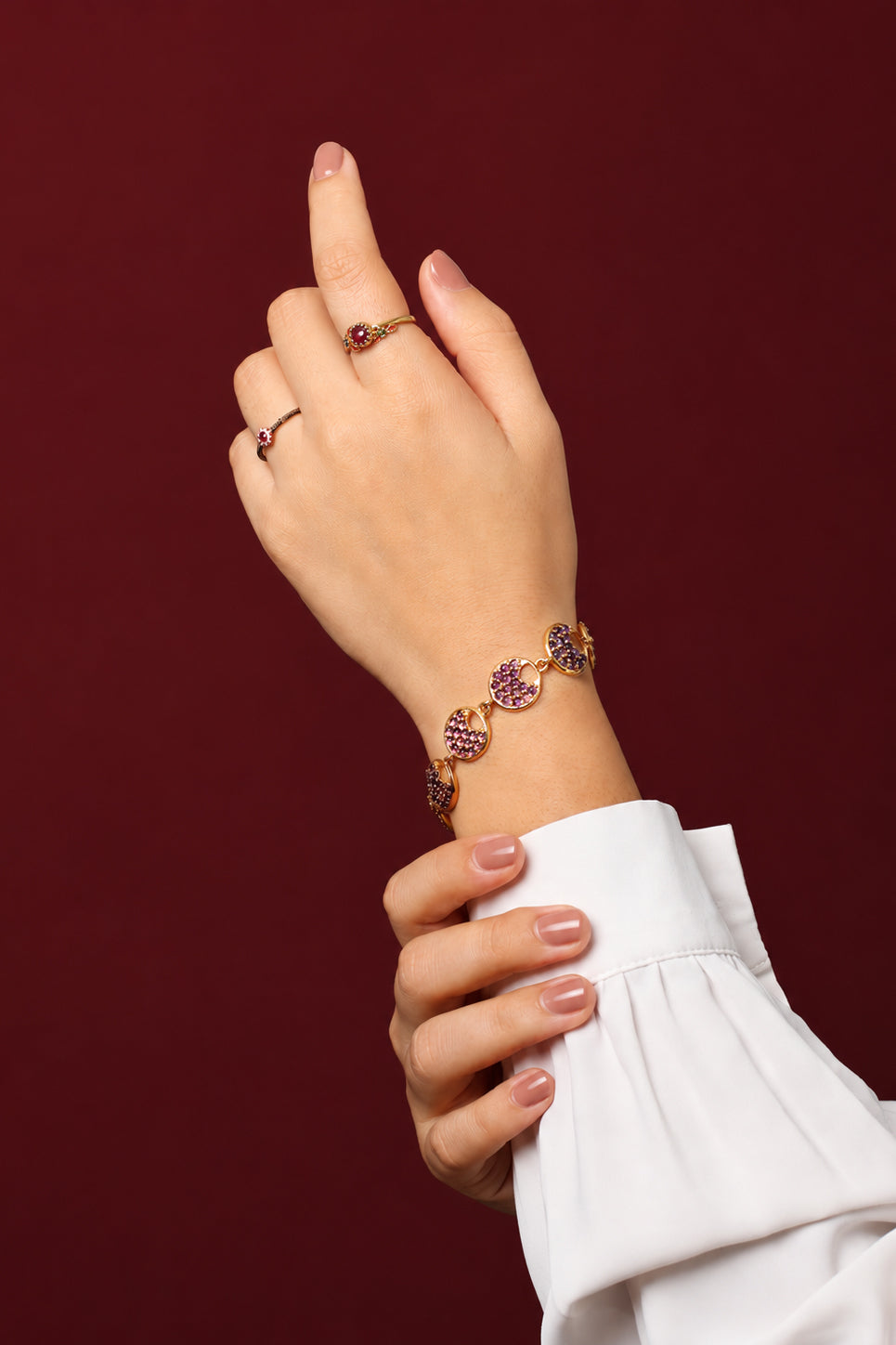 A close-up of a person's hands and wrist wearing jewelry against a dark burgundy background. 