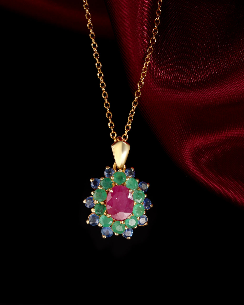 A gold pendant necklace featuring a central oval ruby-red gemstone surrounded by a halo of round green emerald-cut stones and blue round-cut stones, displayed against a dark background with draped red fabric.