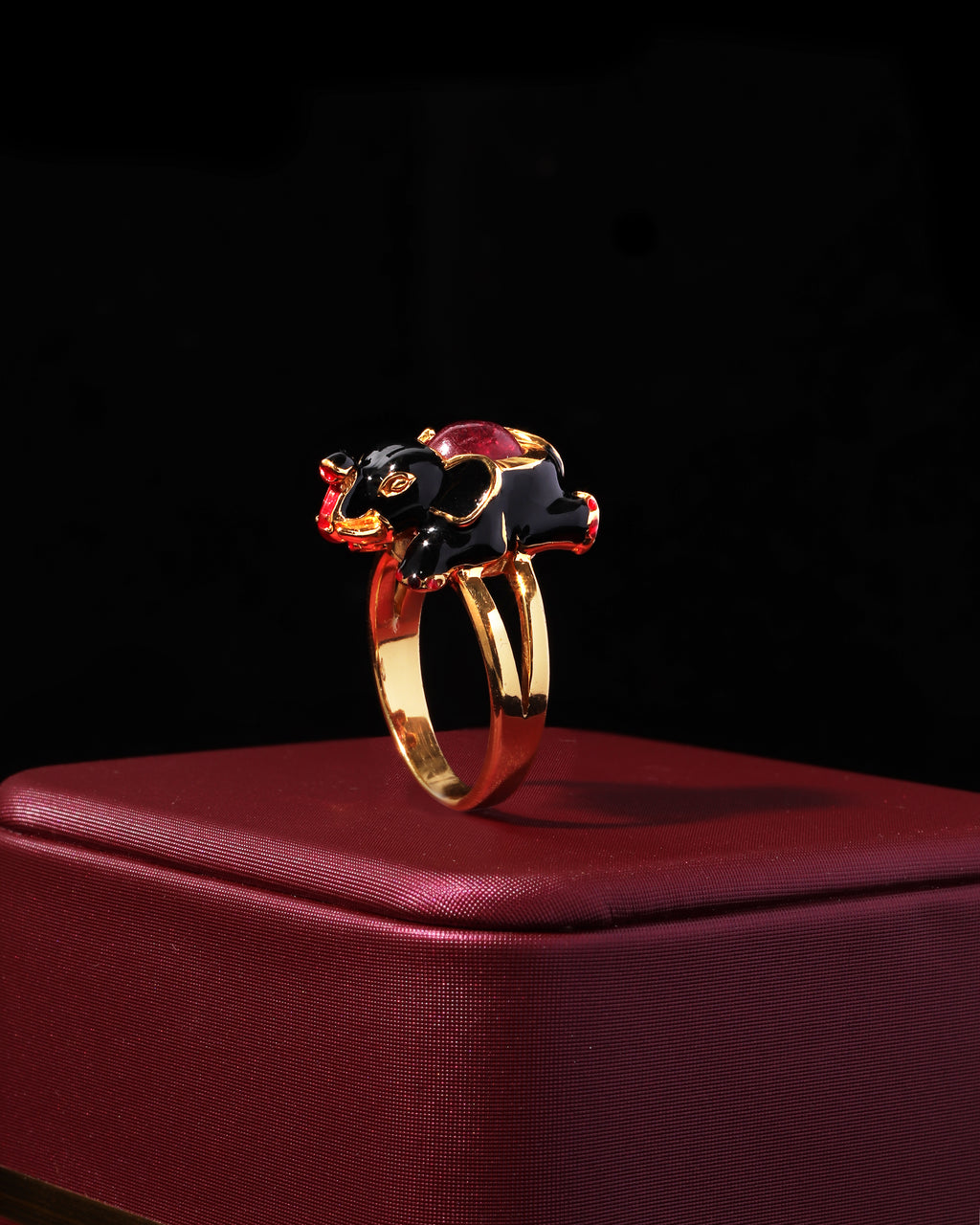 A gold elephant ring with black enamel and red gemstone accents, displayed on a textured dark red box against a black background.