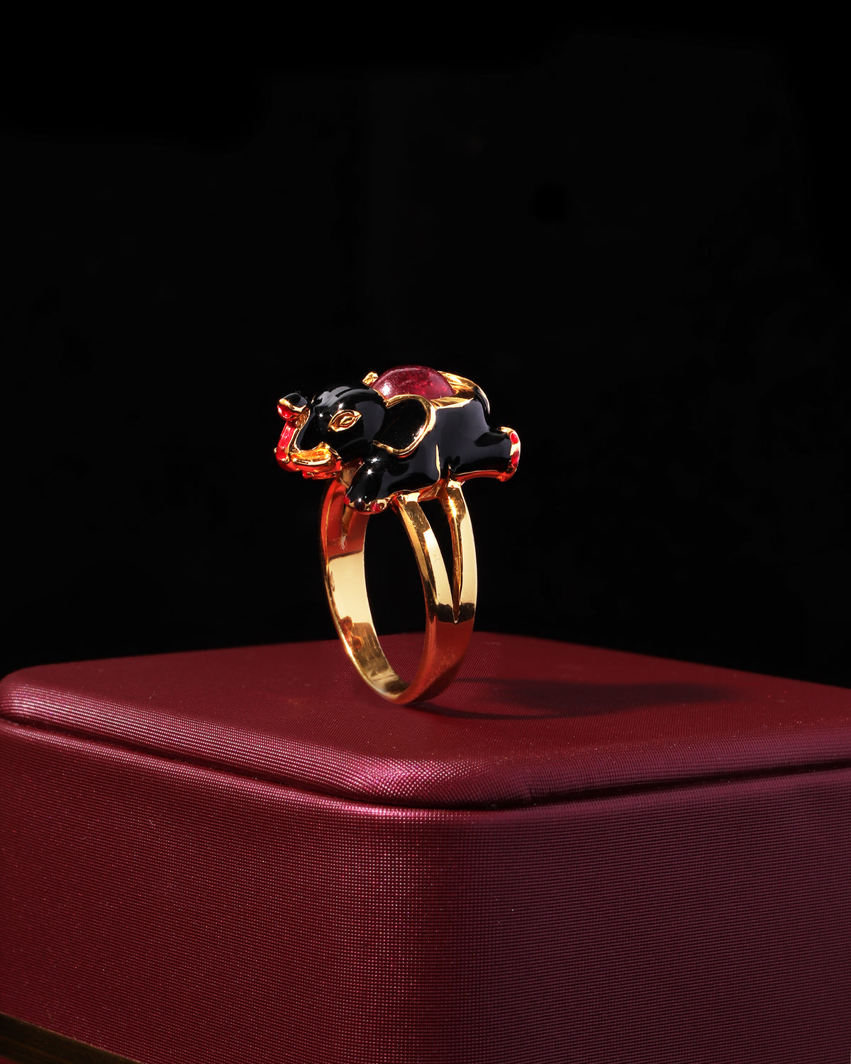 A gold elephant ring with black enamel and red gemstone accents, displayed on a textured dark red box against a black background.