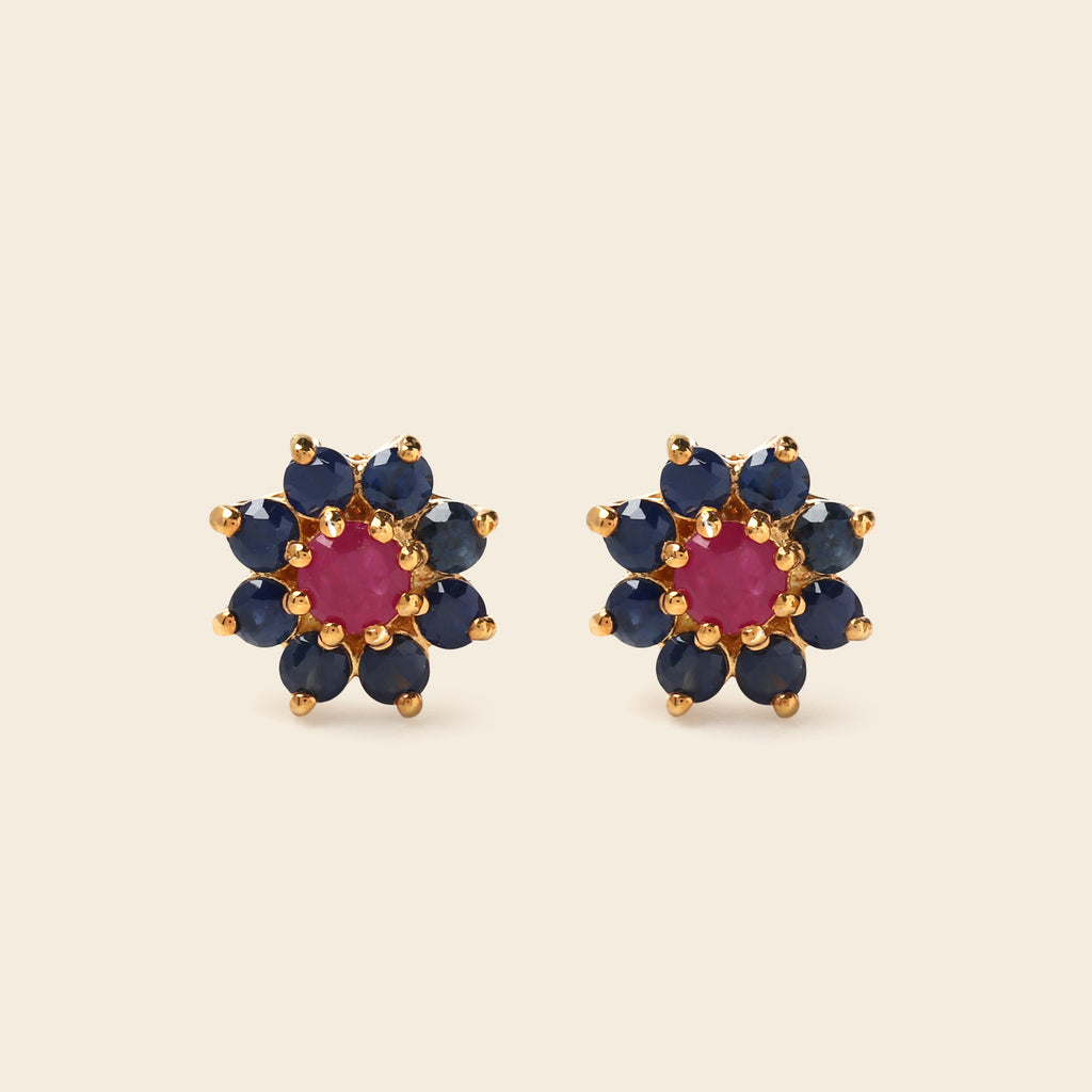 A pair of gold flower-shaped stud earrings featuring a central round pink gemstone surrounded by a halo of dark blue round gemstones, set against a solid light beige background.