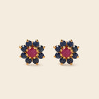 A pair of gold flower-shaped stud earrings featuring a central round pink gemstone surrounded by a halo of dark blue round gemstones, set against a solid light beige background.