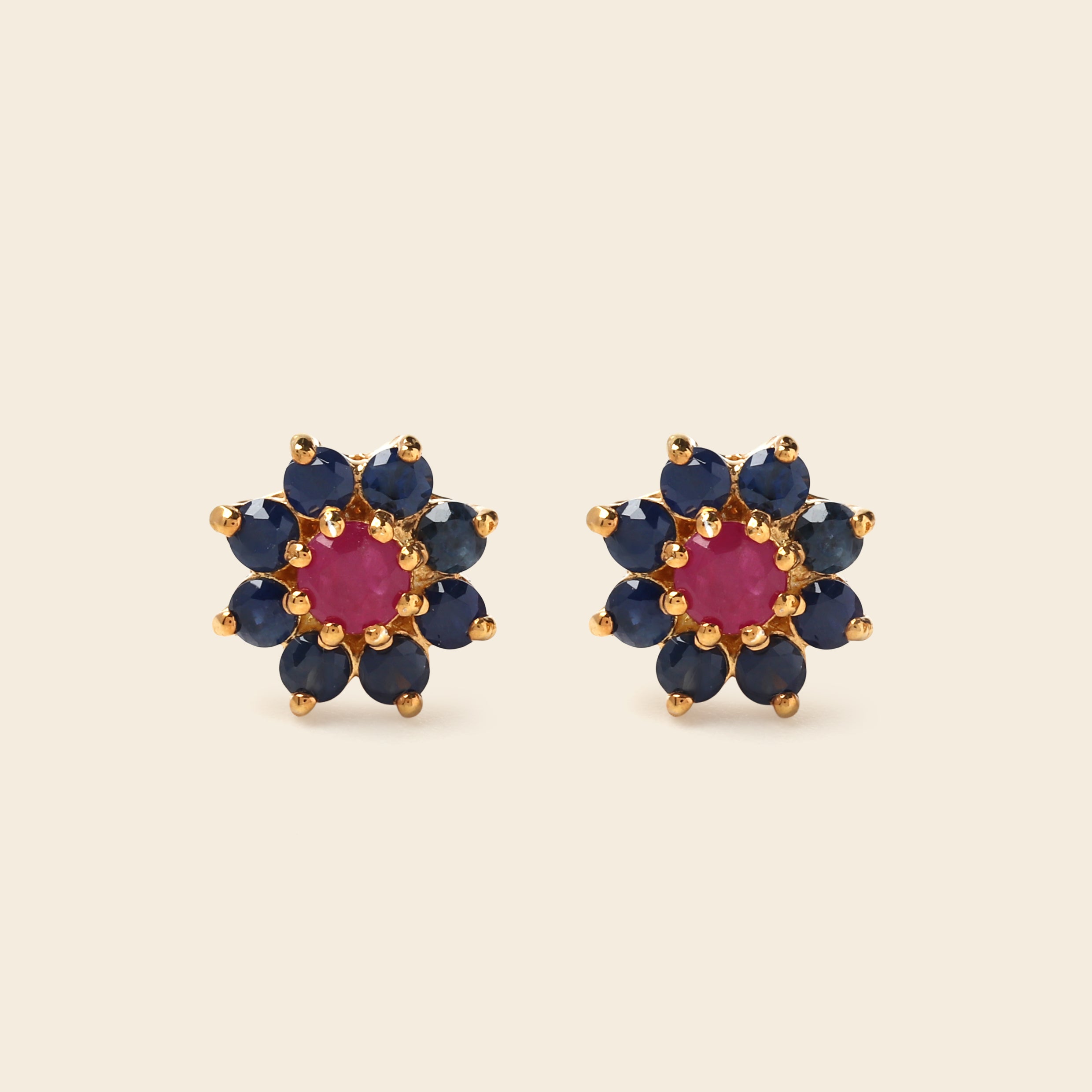 A pair of gold flower-shaped stud earrings featuring a central round pink gemstone surrounded by a halo of dark blue round gemstones, set against a solid light beige background.