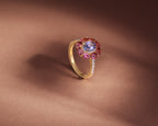 A yellow gold ring with a round cluster setting featuring a central light blue oval gemstone surrounded by pink and red stones, accented with small clear stones on the band, set against a warm brown textured background.