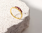 A gold ring with a row of small purple gemstones, displayed on a textured white stone slab against a clean white background.