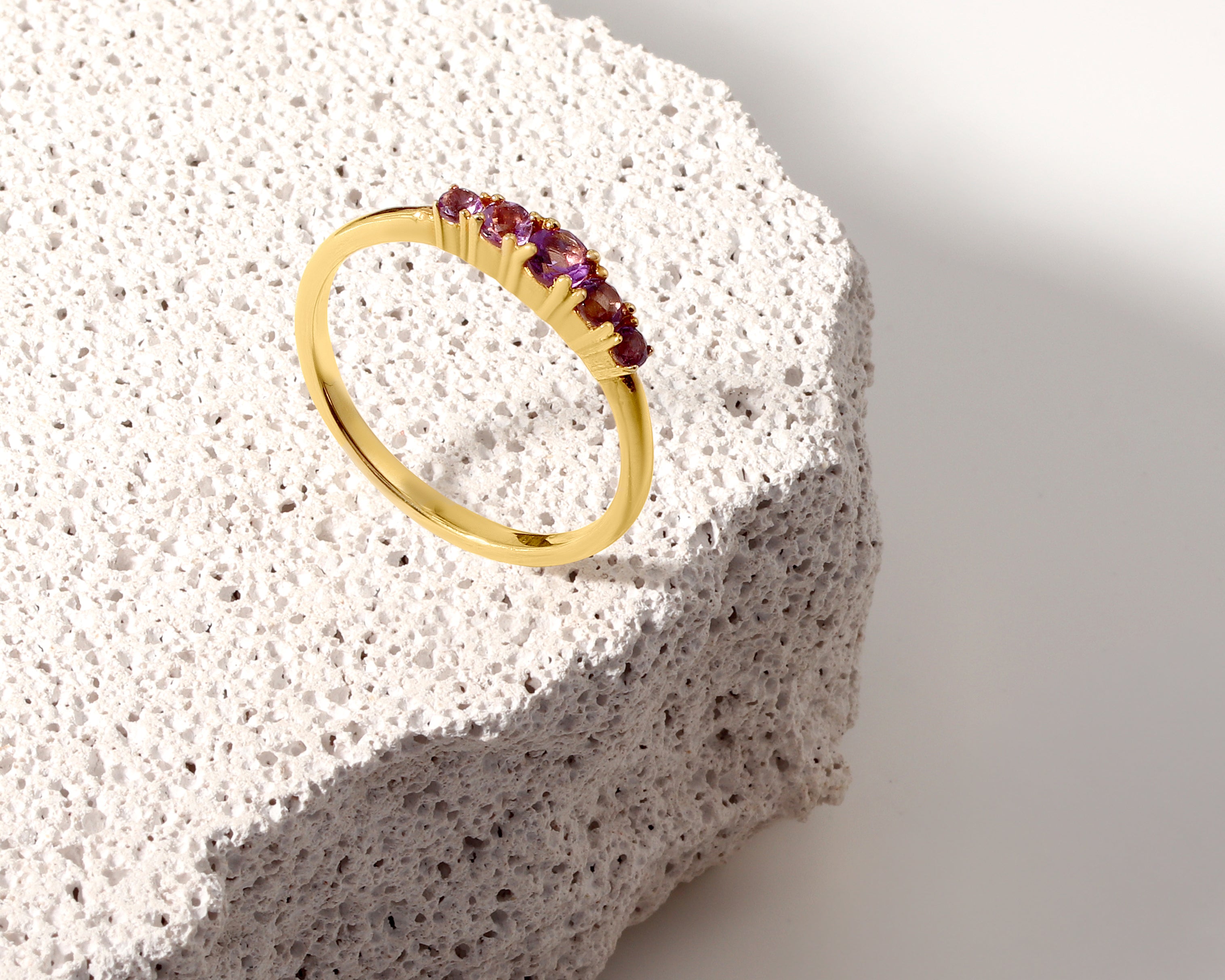 A gold ring with a row of small purple gemstones, displayed on a textured white stone slab against a clean white background.