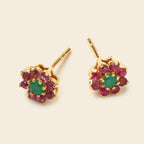 A pair of gold flower-shaped stud earrings featuring a central green emerald-cut stone surrounded by a ring of round pinkish-red gemstones, set against a light beige background.