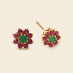 A pair of gold flower-shaped stud earrings featuring a central green emerald-cut stone surrounded by a halo of round pinkish-red ruby stones, shown on a light beige background.