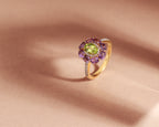 A yellow gold ring with a central green peridot gemstone surrounded by a halo of purple amethyst and small clear accent stones, displayed on a textured beige surface.