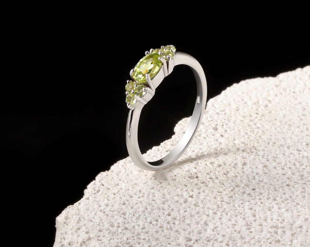 A silver ring with a marquise-cut peridot center stone and small round accent stones, displayed on a textured white stone against a black background.
