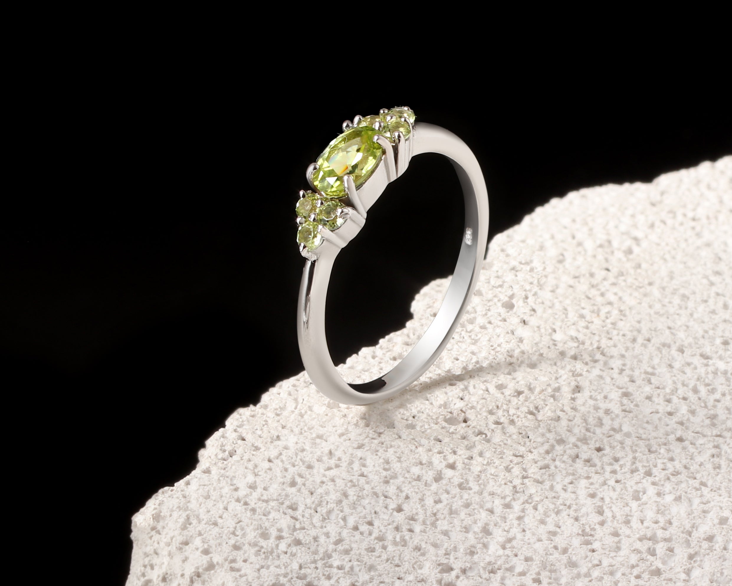 A silver ring with a marquise-cut peridot center stone and small round accent stones, displayed on a textured white stone against a black background.