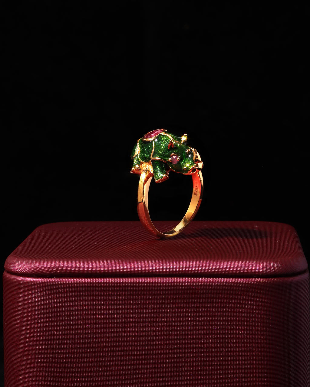 A gold ring with a floral cluster setting featuring green and pink enamel, displayed on a dark red velvet jewelry box against a black background.