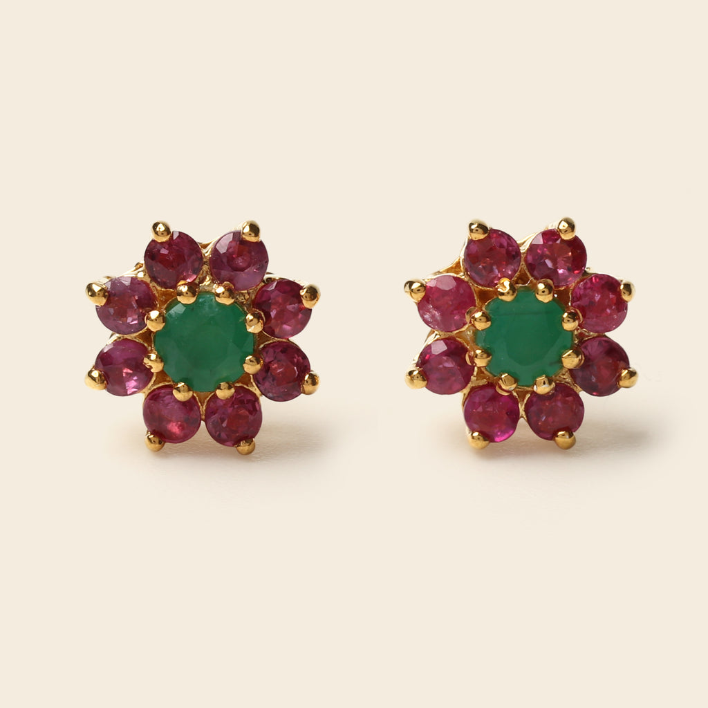 A pair of gold flower-shaped stud earrings featuring a central green emerald-cut stone surrounded by a halo of round pinkish-red ruby-cut stones, set against a light beige background.