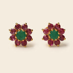 A pair of gold flower-shaped stud earrings featuring a central green emerald-cut stone surrounded by a halo of round pinkish-red ruby-cut stones, set against a light beige background.