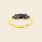A yellow gold ring featuring a central oval dark blue gemstone flanked by four smaller round blue gemstones, set against a plain light beige background.