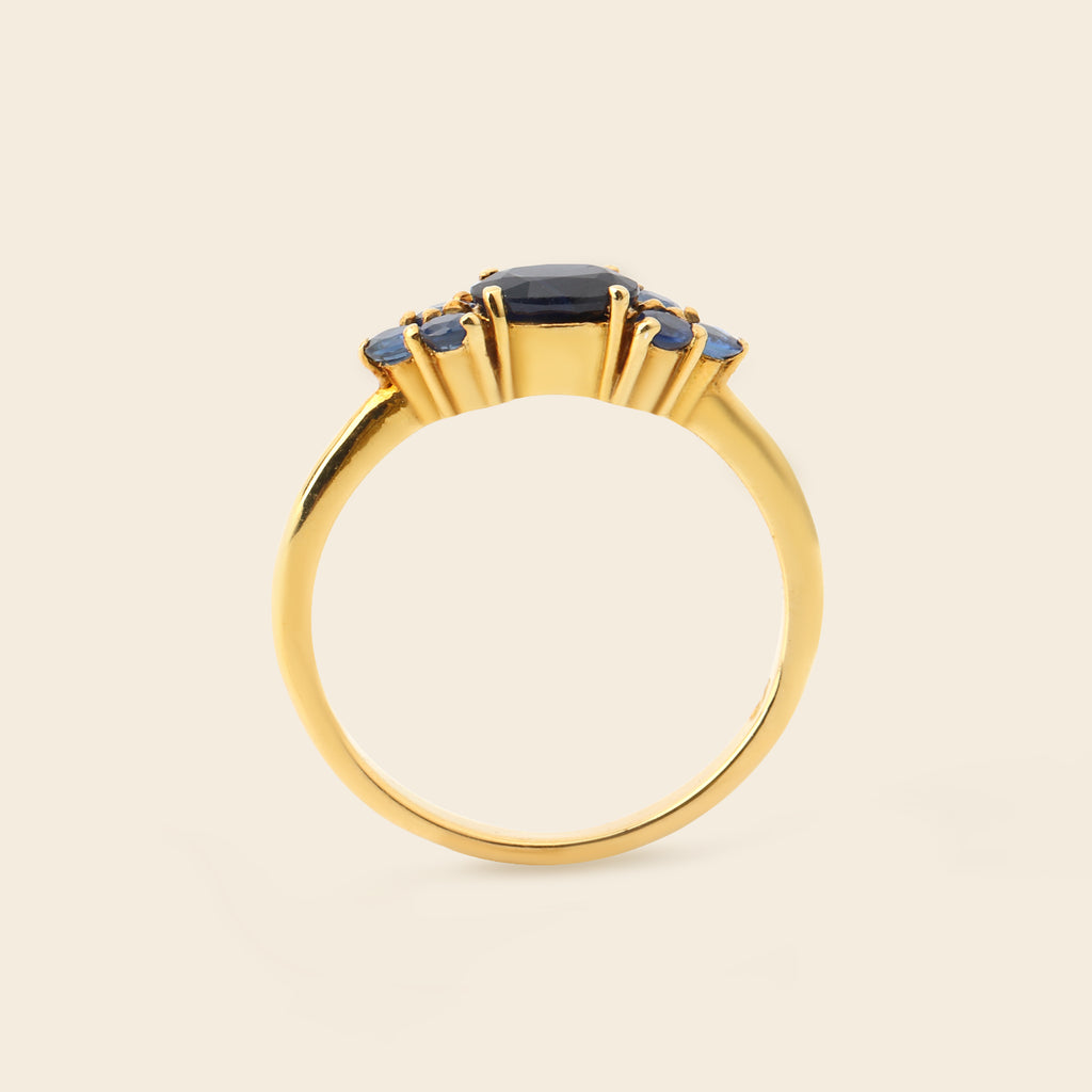 A gold ring featuring a central dark blue oval gemstone and three smaller blue oval gemstones on either side, set against a solid light beige background.